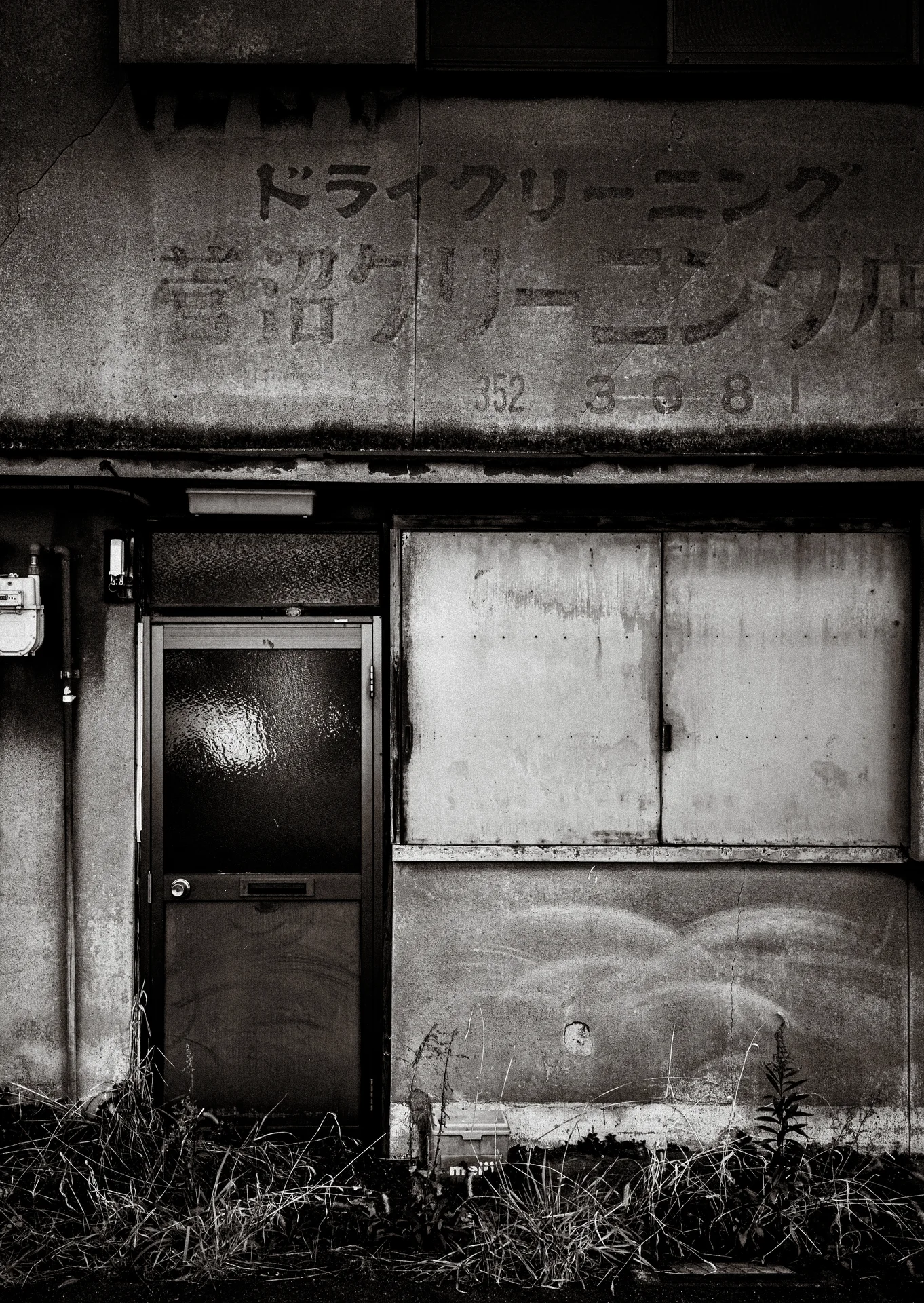 Faded lettering marks a weathered facade above a narrow door, boarded window, and weeds.