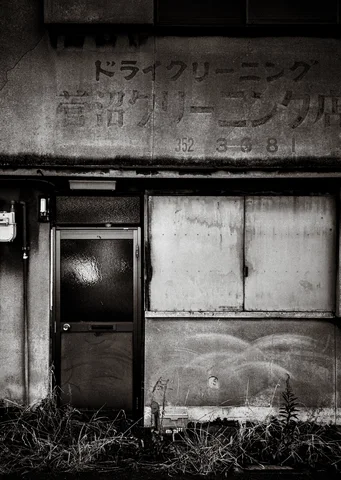 Faded lettering marks a weathered facade above a narrow door, boarded window, and weeds.
