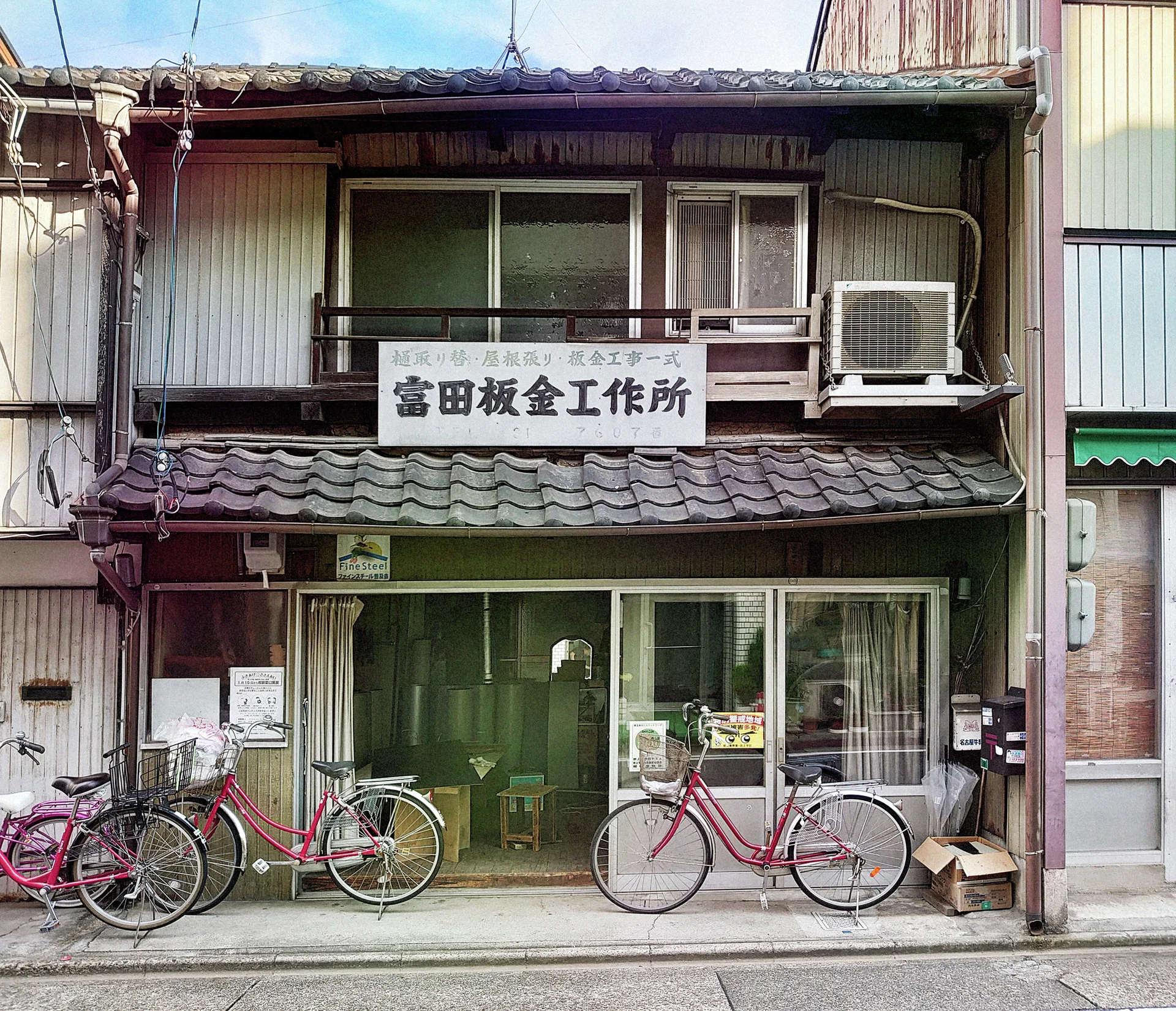 Several bicycles line a weathered storefront beneath tiled eaves, an upstairs window, and an air conditioner.