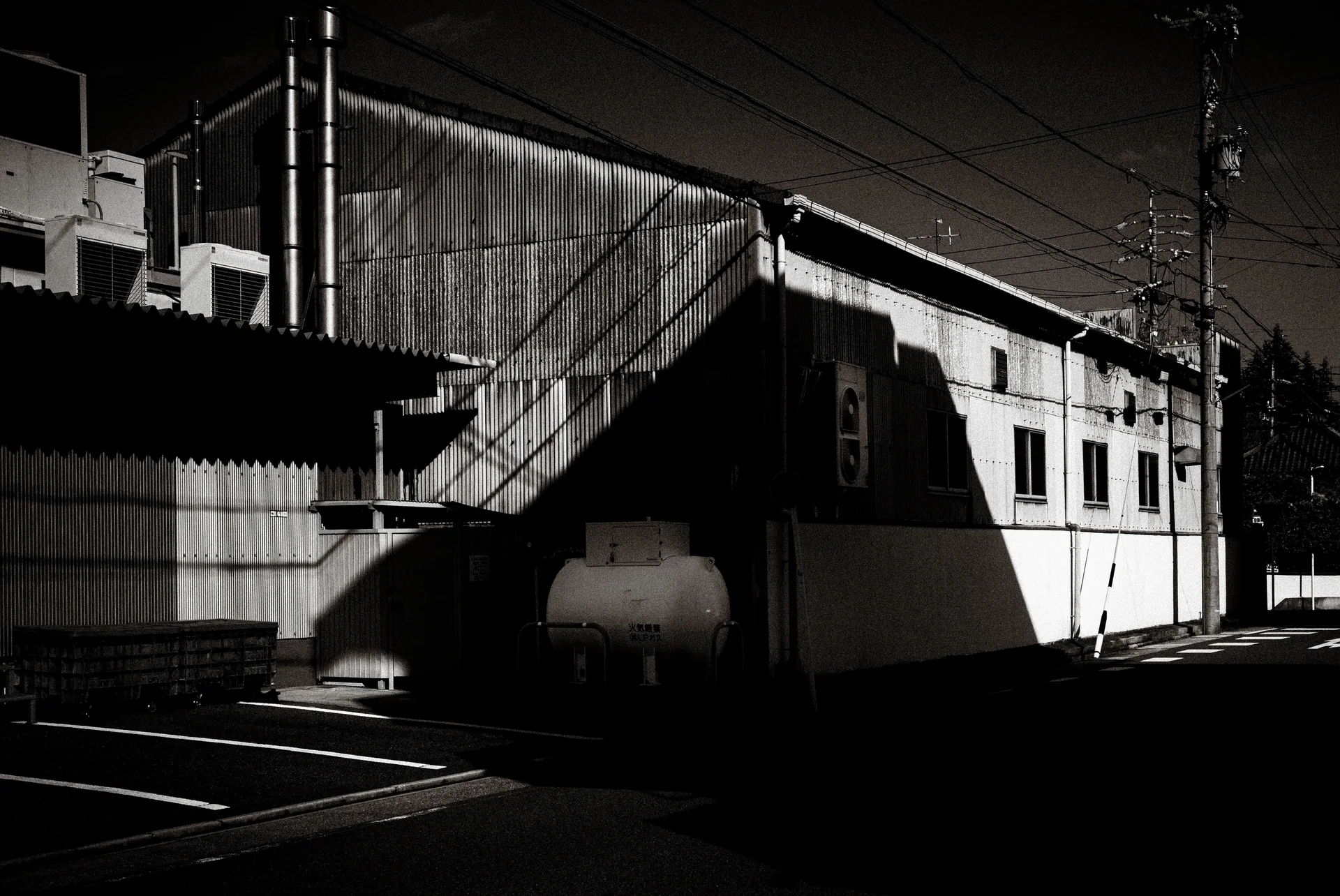 Sharp diagonal shadows cross a long corrugated industrial building beneath overhead wires in black and white.