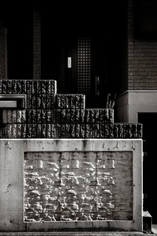 A patterned concrete wall fronts a shadowed entrance behind stepped dark blocks in black and white.