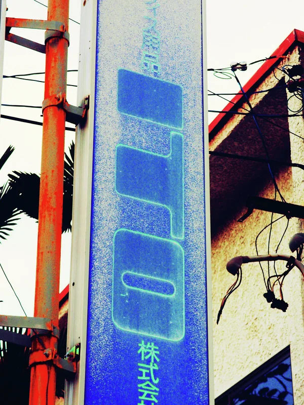 Faded large blue lettering runs down a narrow vertical sign beside orange pipes and pale walls.