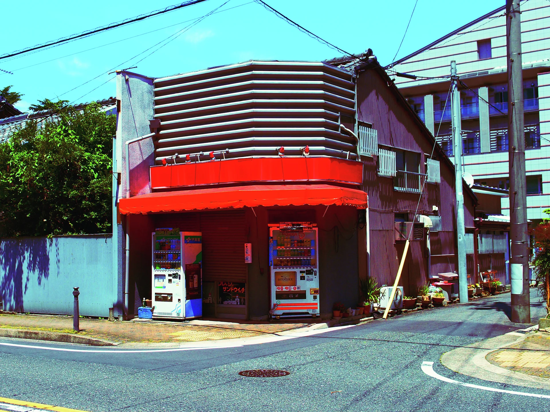 A red corner awning shelters two vending machines beneath curved horizontal metal panels on an older building.