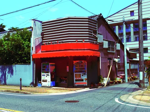 A red corner awning shelters two vending machines beneath curved horizontal metal panels on an older building.