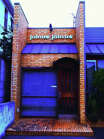 A narrow brick storefront has a blue shutter, arched dark doorway, and small lettering above a blue awning.