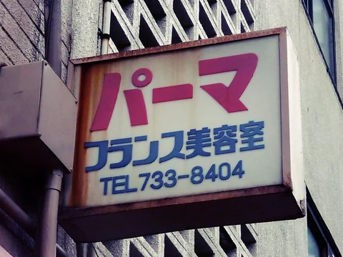 A weathered projecting salon sign displays large pink Japanese characters above smaller blue lettering.