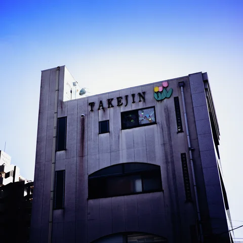 Dark TAKEJIN letters and a small colorful emblem sit atop a tall pale facade with recessed windows.