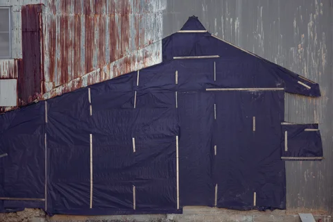 Purple sheeting patches a house-shaped section of a weathered corrugated metal wall.