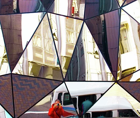 Triangular mirrors reflect buildings, a white van and a person in red holding a camera.