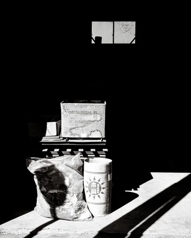 Bags and a bucket catch sunlight at the entrance of a dark workshop in black and white.