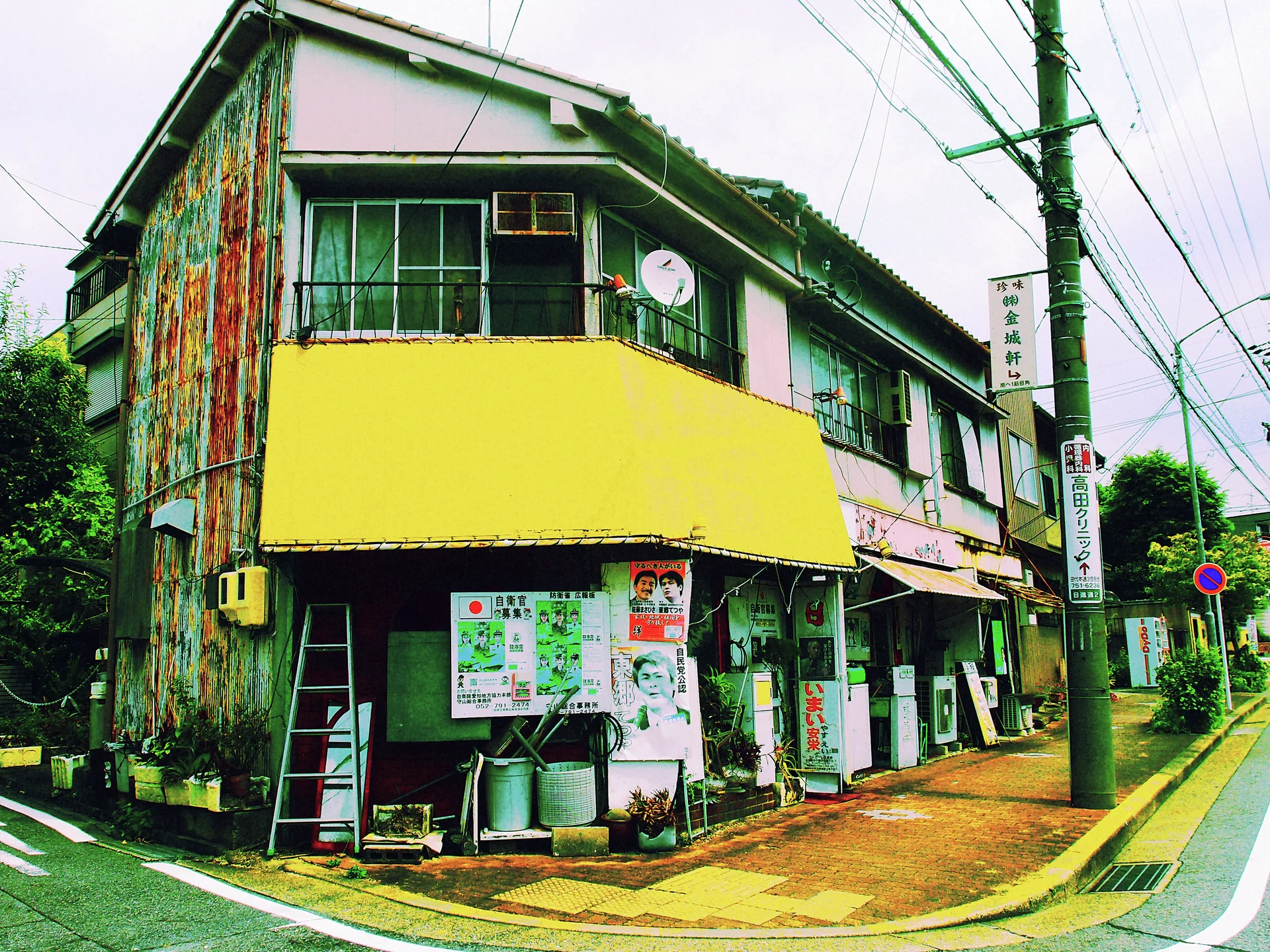A weathered two-story corner building has a broad yellow facade panel, upper windows, and posters around its entrance.