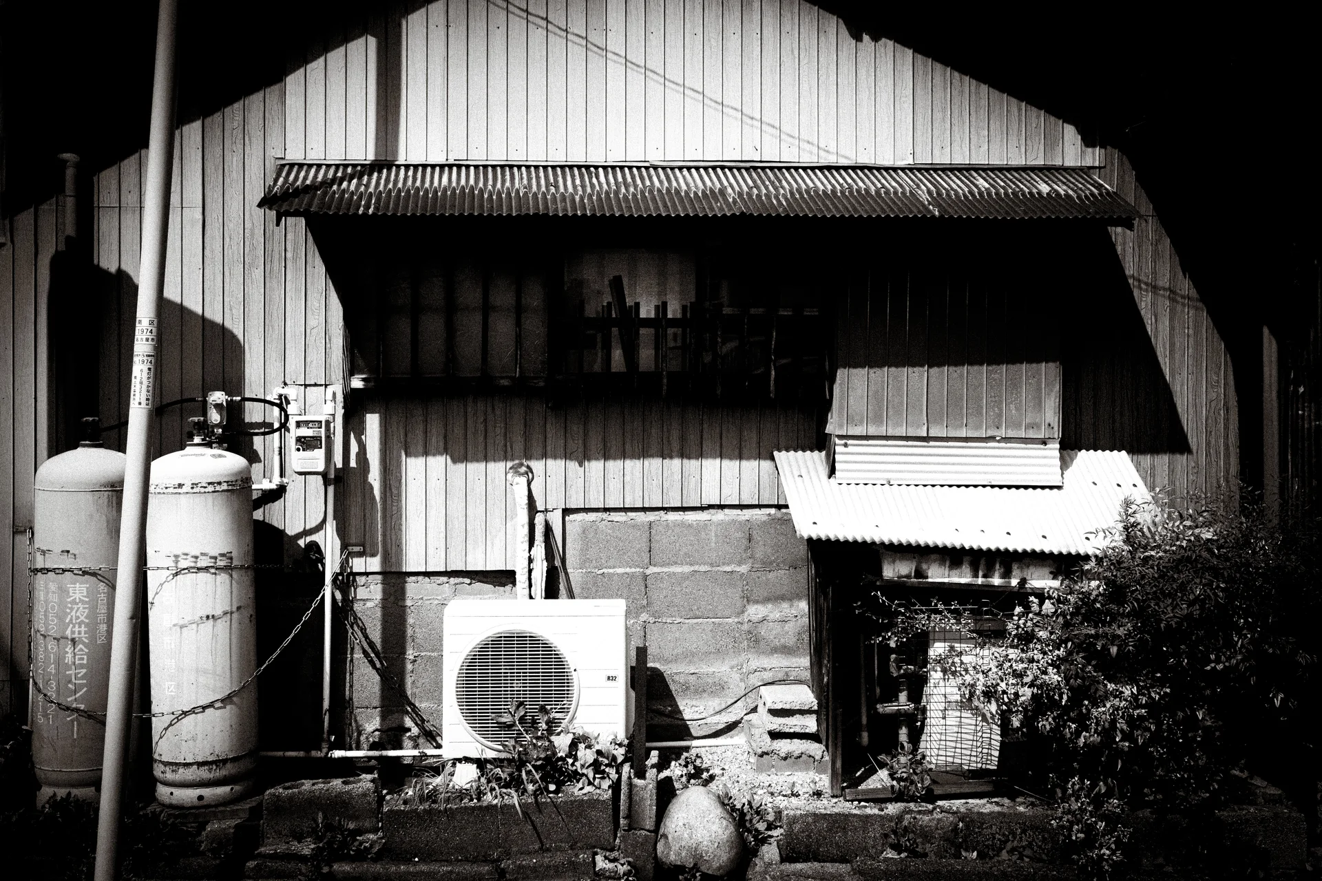 A cylindrical tank and air conditioner stand outside a corrugated house with deep awning shadows in black and white.