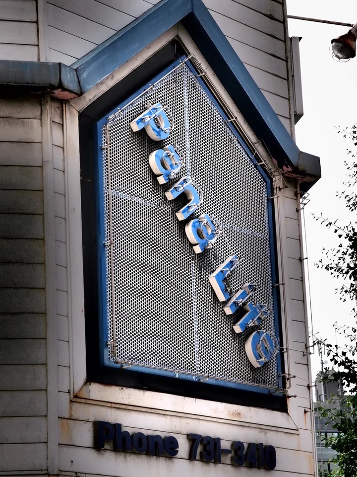 Angled pale letters descend across a dark mesh sign framed by a pointed blue border.