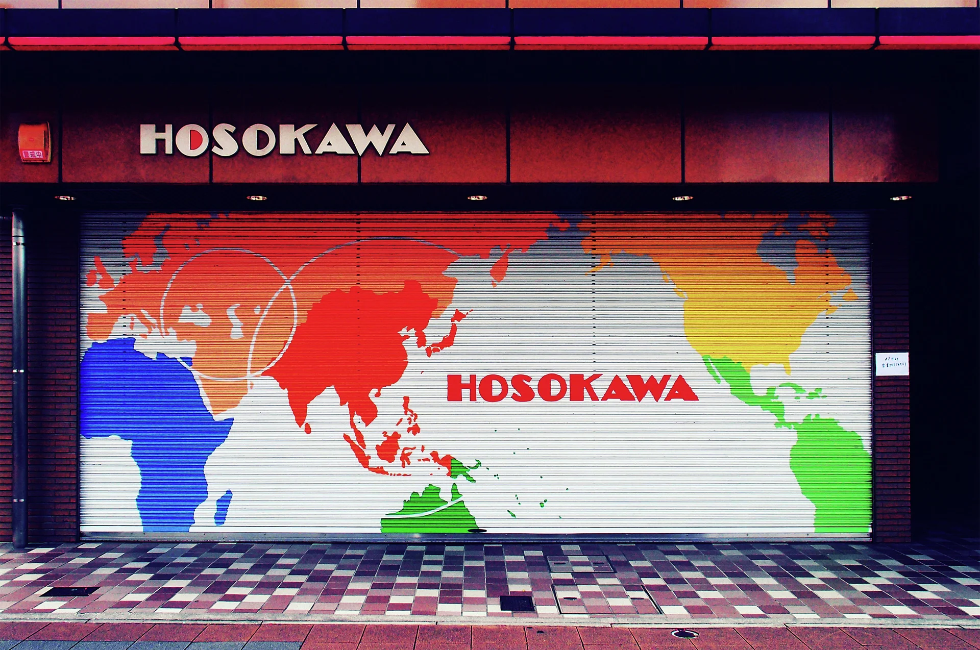 A colorful world map covers a closed storefront shutter above a checker-patterned tiled pavement.