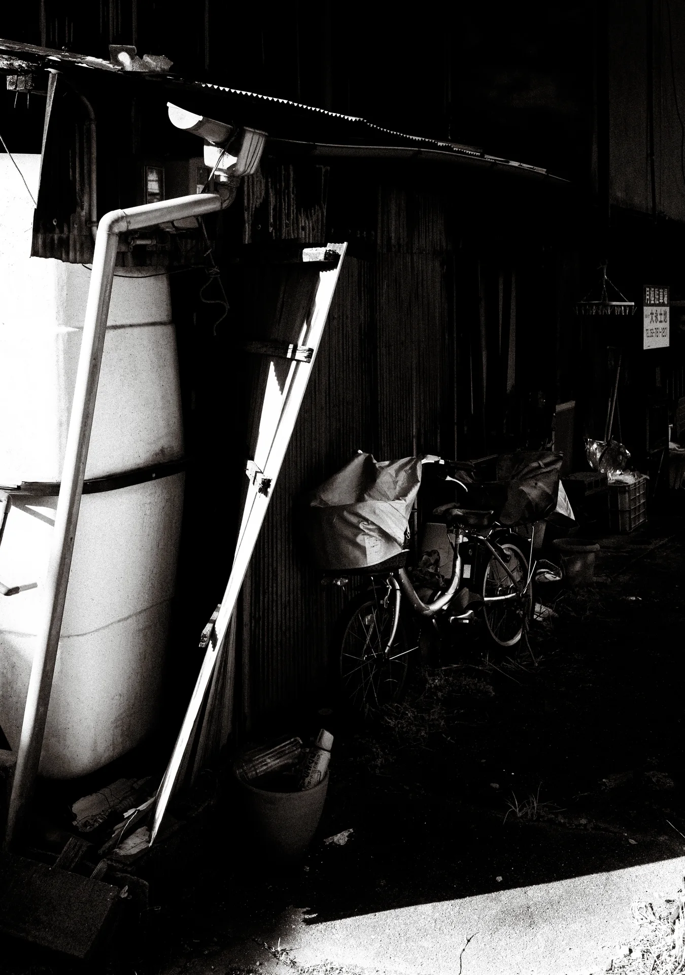 Two long ladders lean beside a large pale panel in a dark cluttered space in black and white.