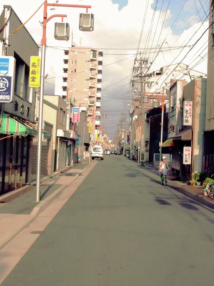 A quiet narrow street extends between shops and tall buildings beneath dense overhead utility wires.