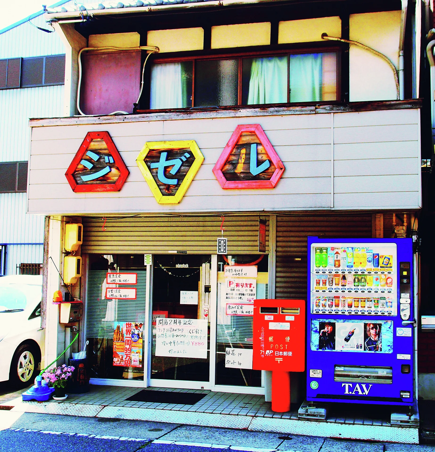 Three colorful geometric letter panels mark a small storefront beside a red post box and blue vending machine.