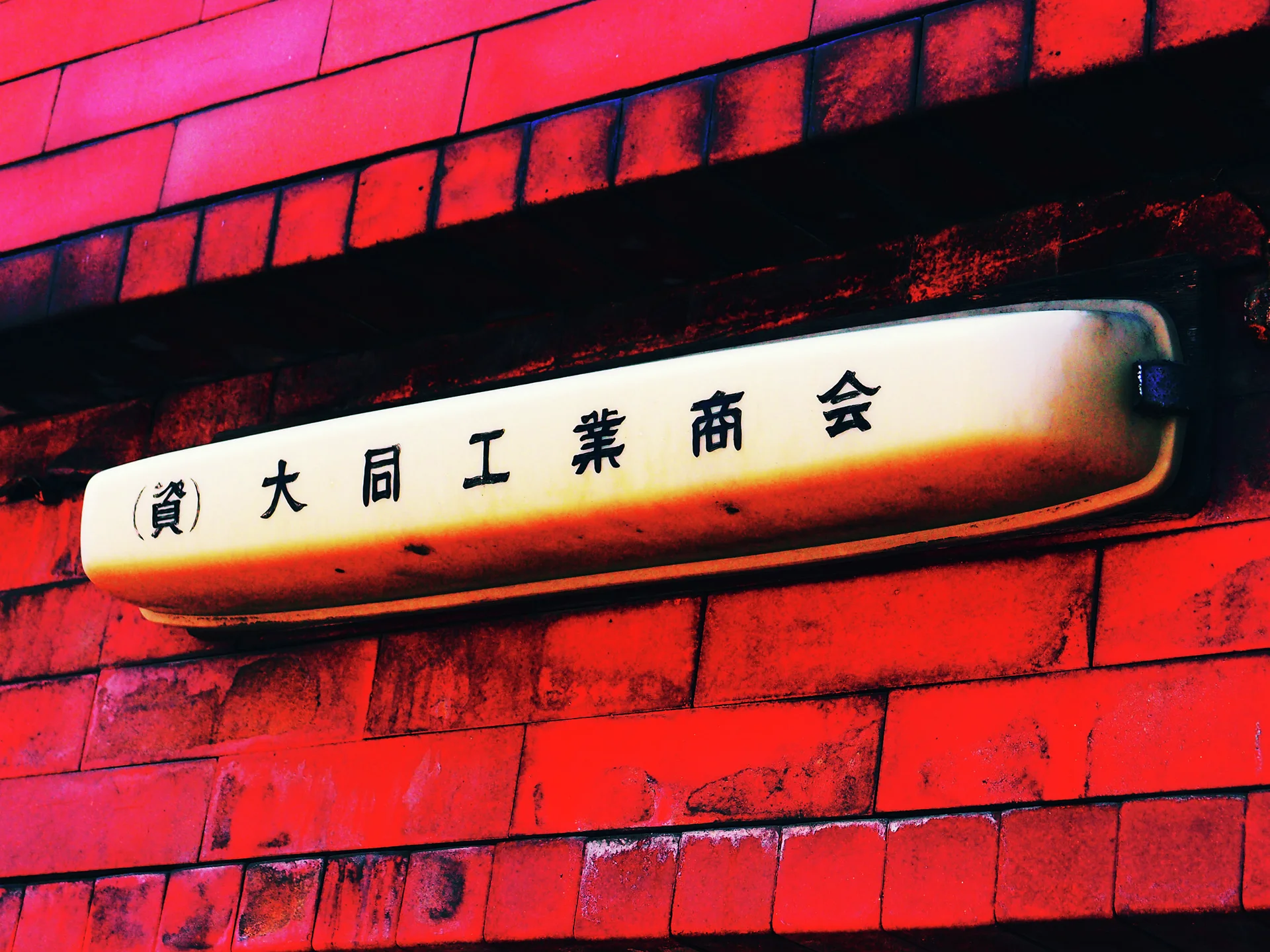 A narrow cream sign with black Japanese lettering projects from a vivid red brick wall.