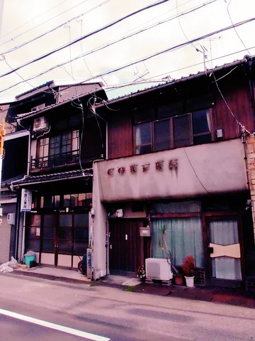 Two narrow weathered buildings stand beneath overhead wires with small plants beside their entrances.
