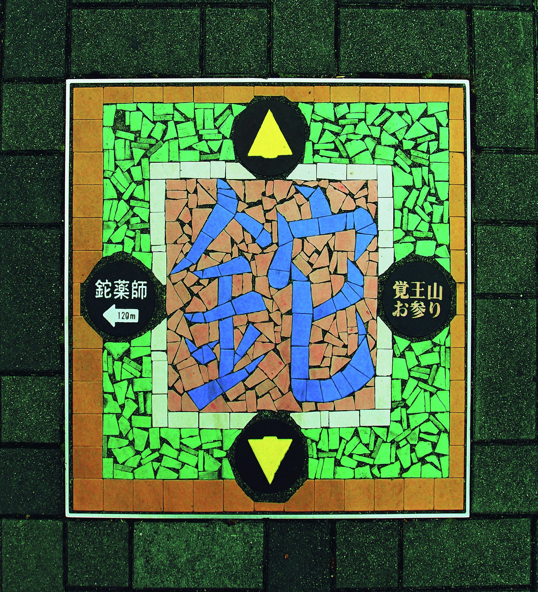 A colorful mosaic panel with blue Japanese characters and yellow arrows is set into a gray block wall.