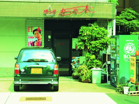 A green compact car is parked outside a dark entrance bordered by potted plants and a vending machine.