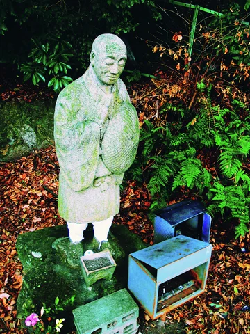A standing robed stone figure holds a rounded object beside small boxes amid ferns and fallen leaves.
