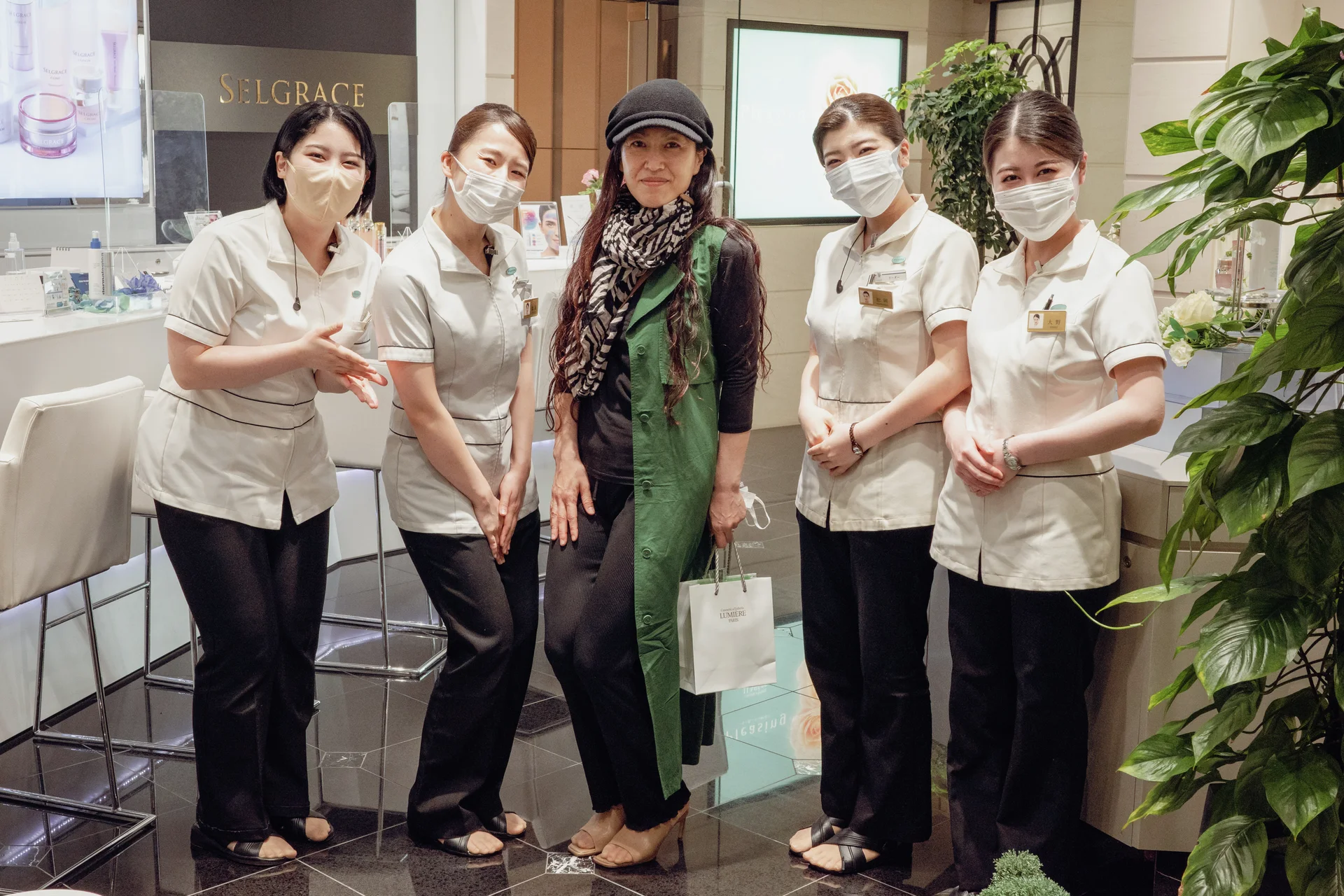 Five adults pose indoors beside leafy plants, four wearing pale uniforms and face masks.