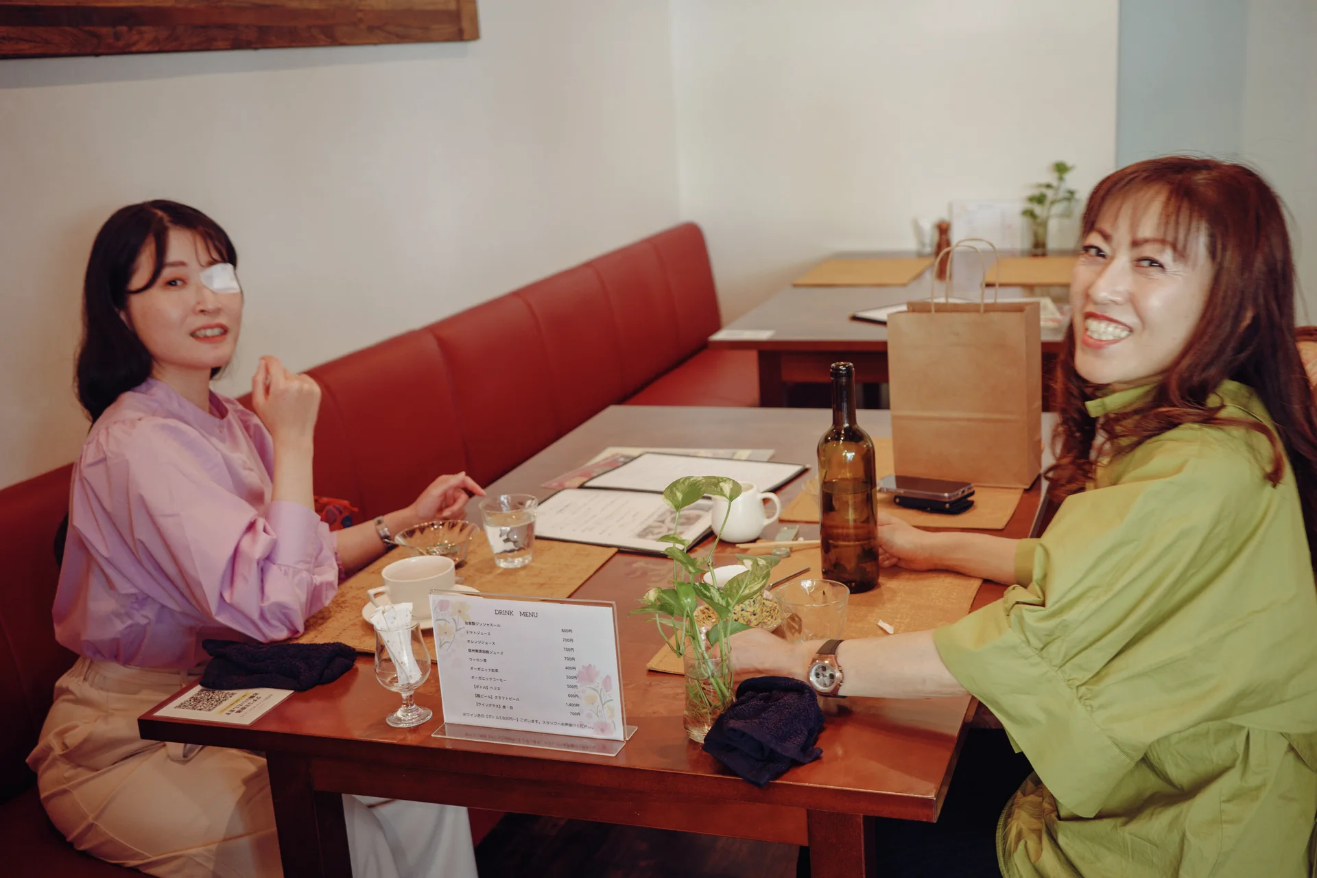 Two women sit across a wooden restaurant table holding dishes, glasses, and bottles beside red bench seating.