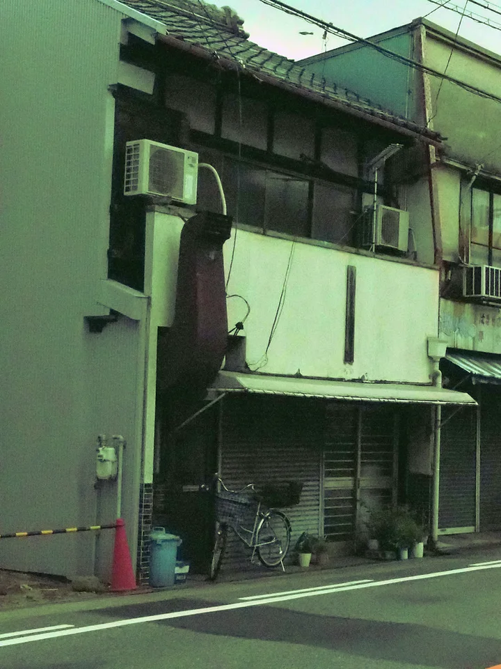 A long metal exhaust duct climbs a pale building facade above a shuttered frontage and parked bicycle.