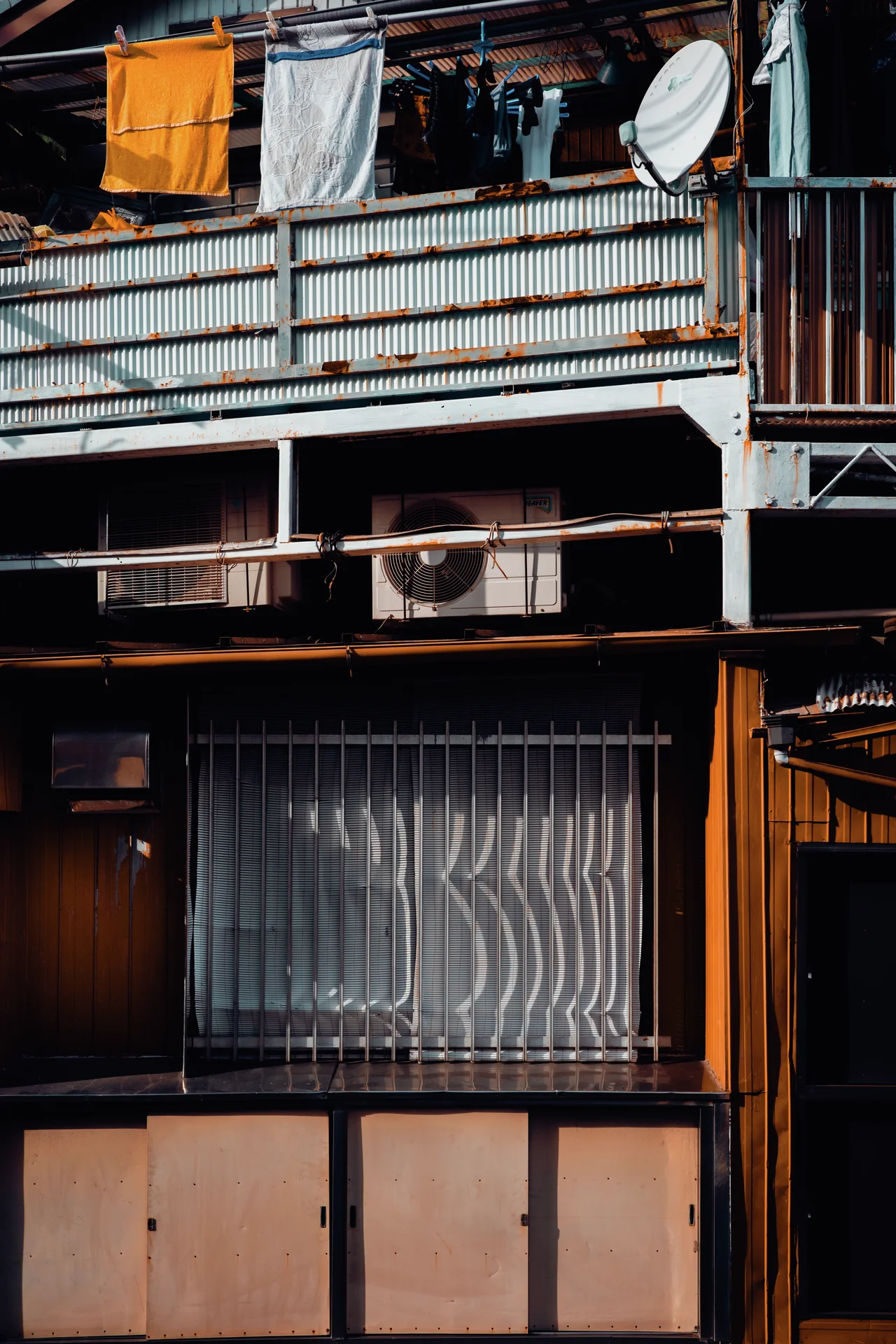 Laundry hangs above a corrugated balcony railing over a barred lower window and wood-paneled facade.