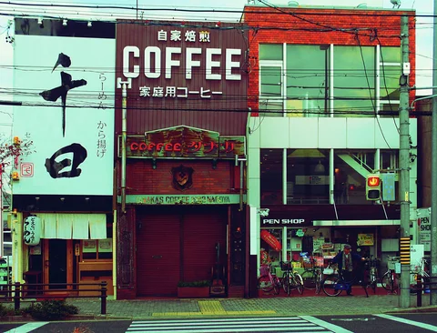 Large COFFEE lettering crowns a narrow brown storefront beside pale green buildings and a striped pedestrian crossing.