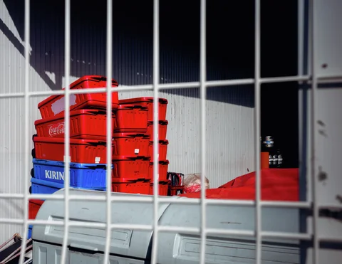 Stacks of red and blue drinks crates sit behind a white metal grille against corrugated siding.