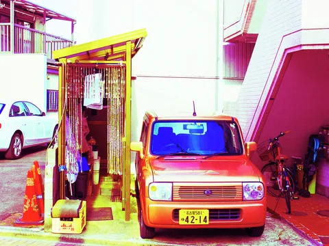 A small orange car parks beside a yellow-roofed outdoor enclosure and a bicycle beneath an exterior staircase.