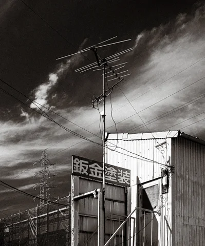 An antenna and overhead wires rise above a corrugated building beneath streaked clouds in black and white.
