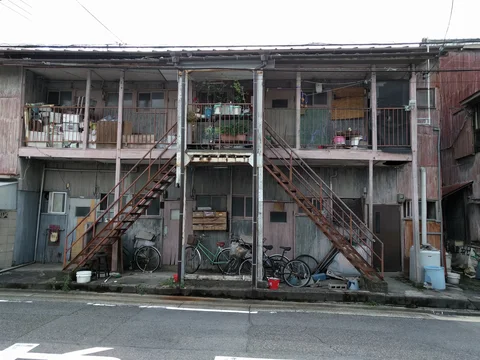 Rust-colored twin staircases flank a central balcony above bicycles outside an aging two-story apartment building.