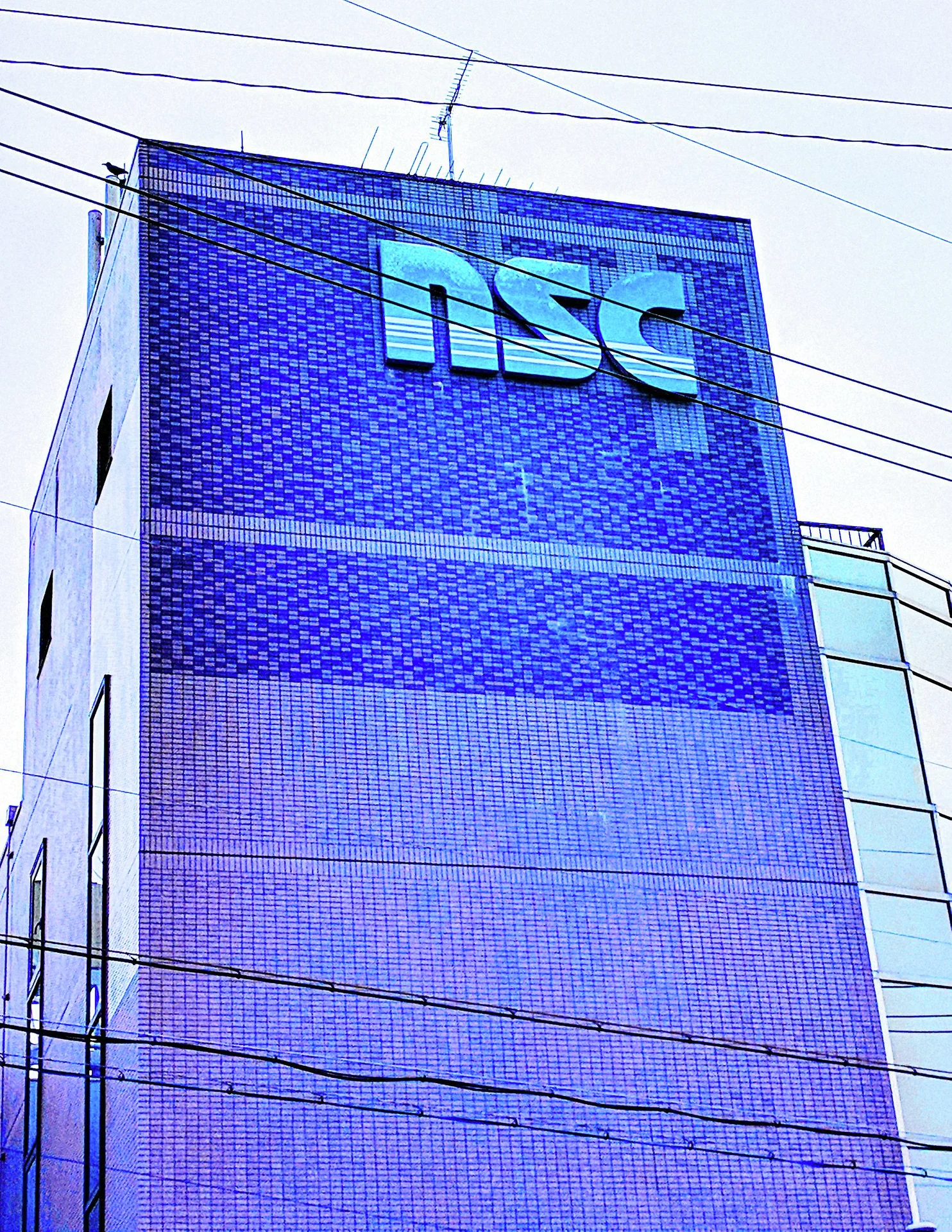 Large turquoise lettering marks the upper corner of a tall blue-tiled building crossed by utility wires.