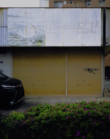 Yellow metal shutters sit beneath a faded white sign behind a hedge with small purple flowers.