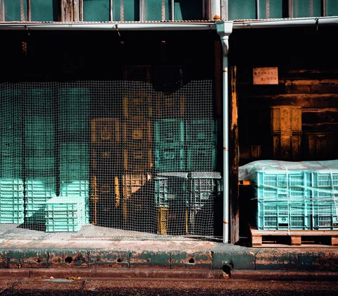 Turquoise plastic crates fill a dim storage frontage behind mesh screening and weathered wooden panels.