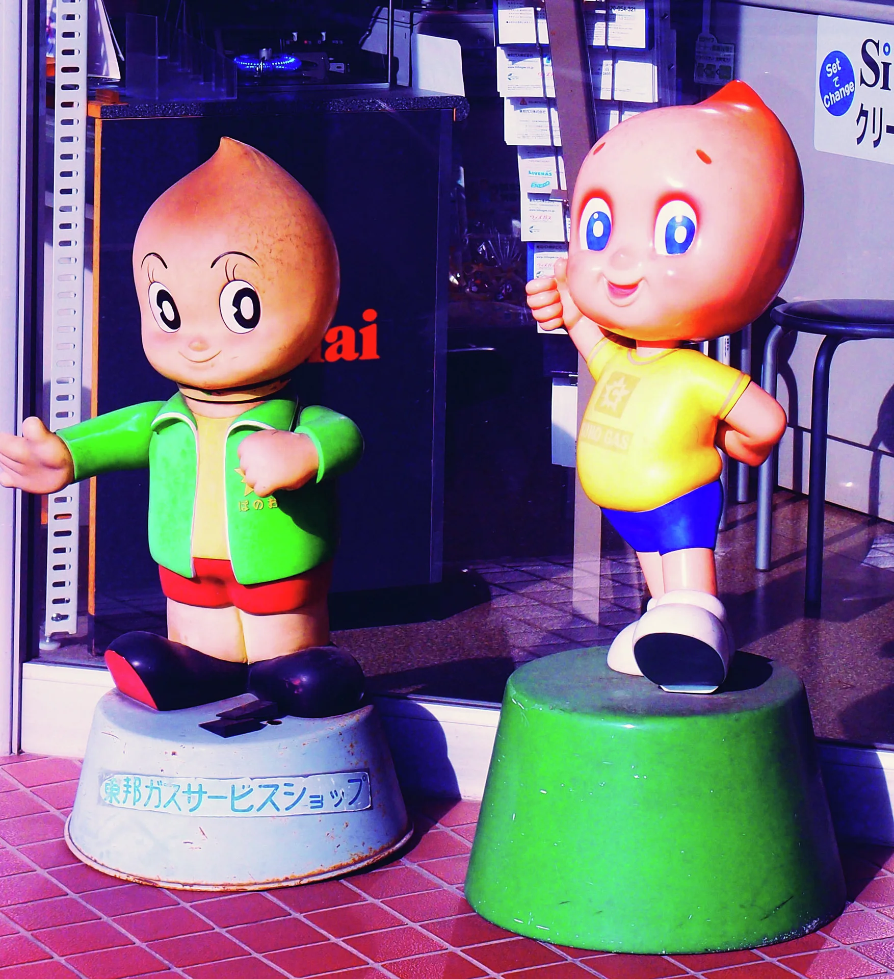 Two smiling cartoon child statues wear green and yellow outfits on round bases outside a shop.