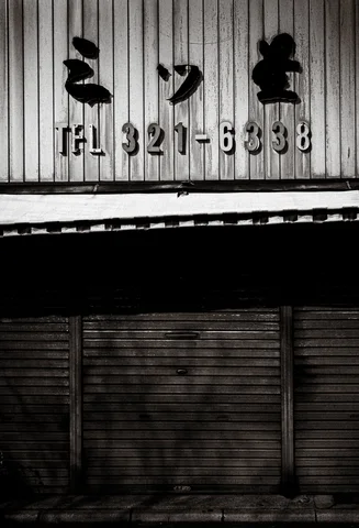 Dark raised characters and telephone numbers appear above closed corrugated shutters in monochrome.