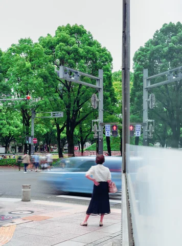 A woman waits beside a tree-lined road as a blurred blue car passes across reflected glass.