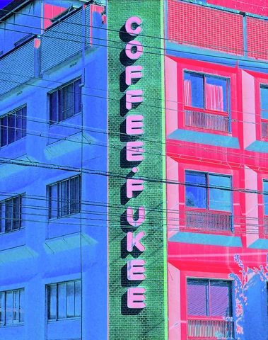 Vertical pale coffee lettering spans a green strip between vivid blue and pink building walls.