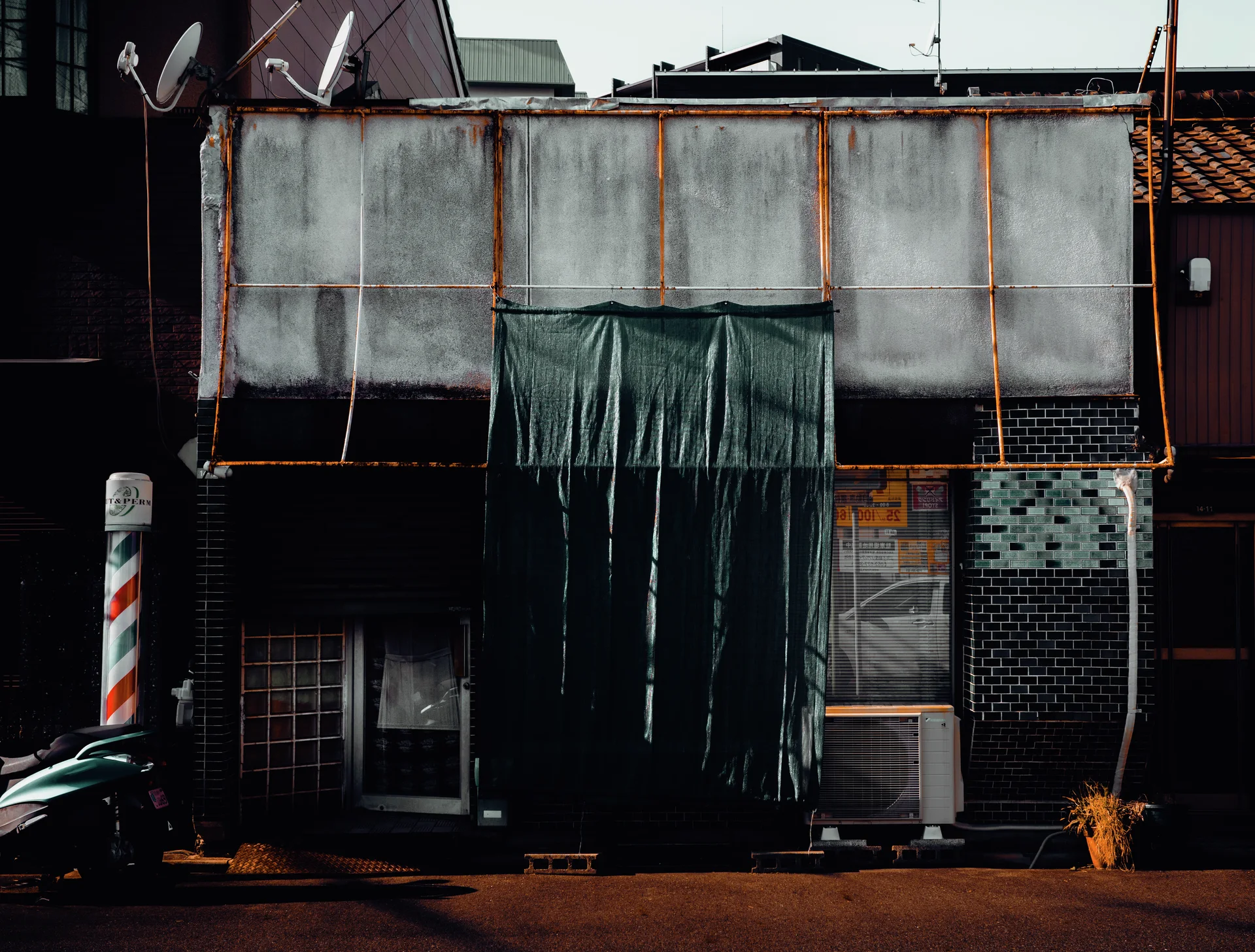 Dark sheeting covers the center of a weathered storefront beside a barber pole and parked scooter.