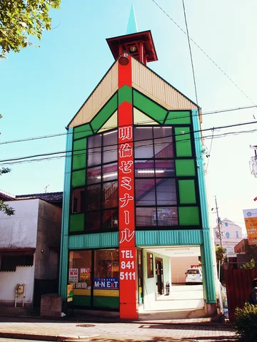A narrow pointed building has green window frames and a tall red vertical sign beneath a small tower.