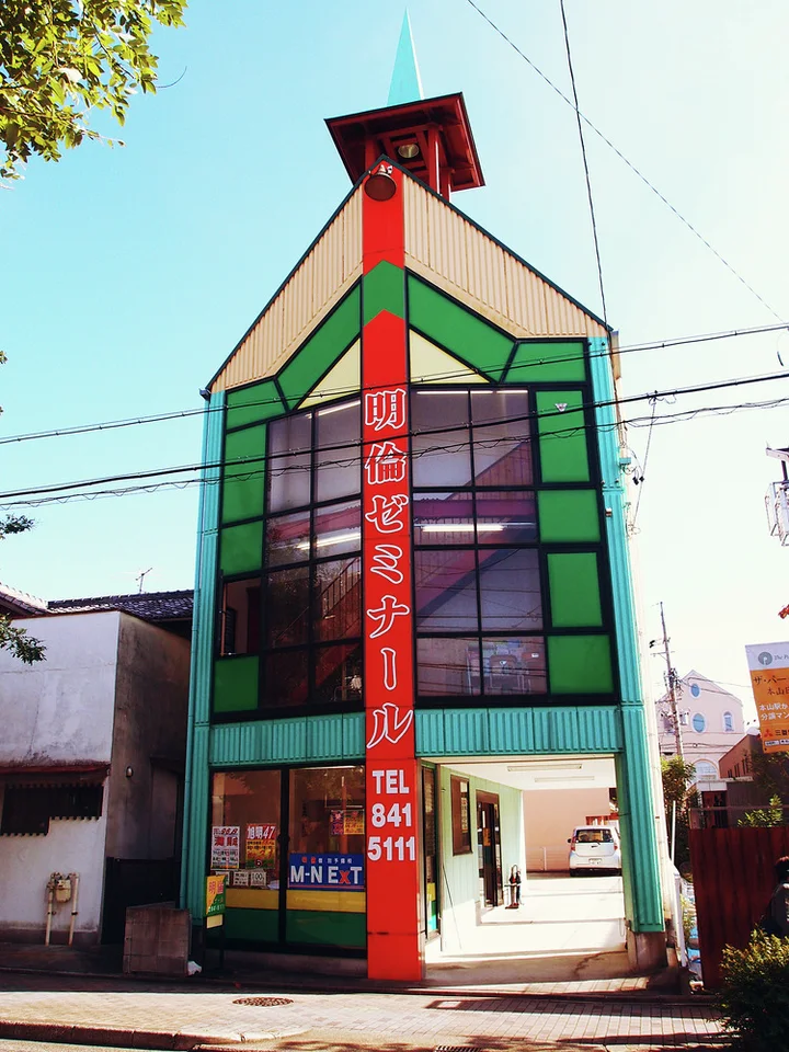 A narrow pointed building has green window frames and a tall red vertical sign beneath a small tower.