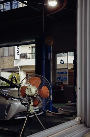 An orange-bladed floor fan stands beside a car inside an open workshop.