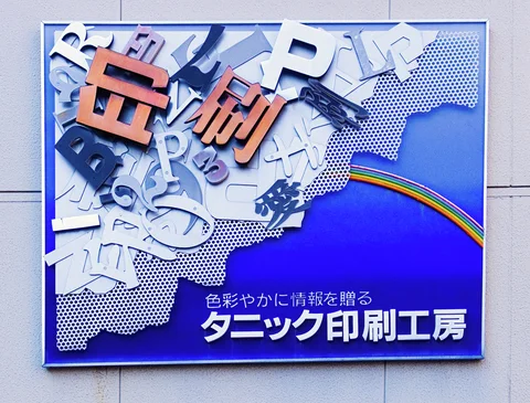 A blue printing sign combines scattered raised letters, orange characters, and a narrow rainbow stripe.