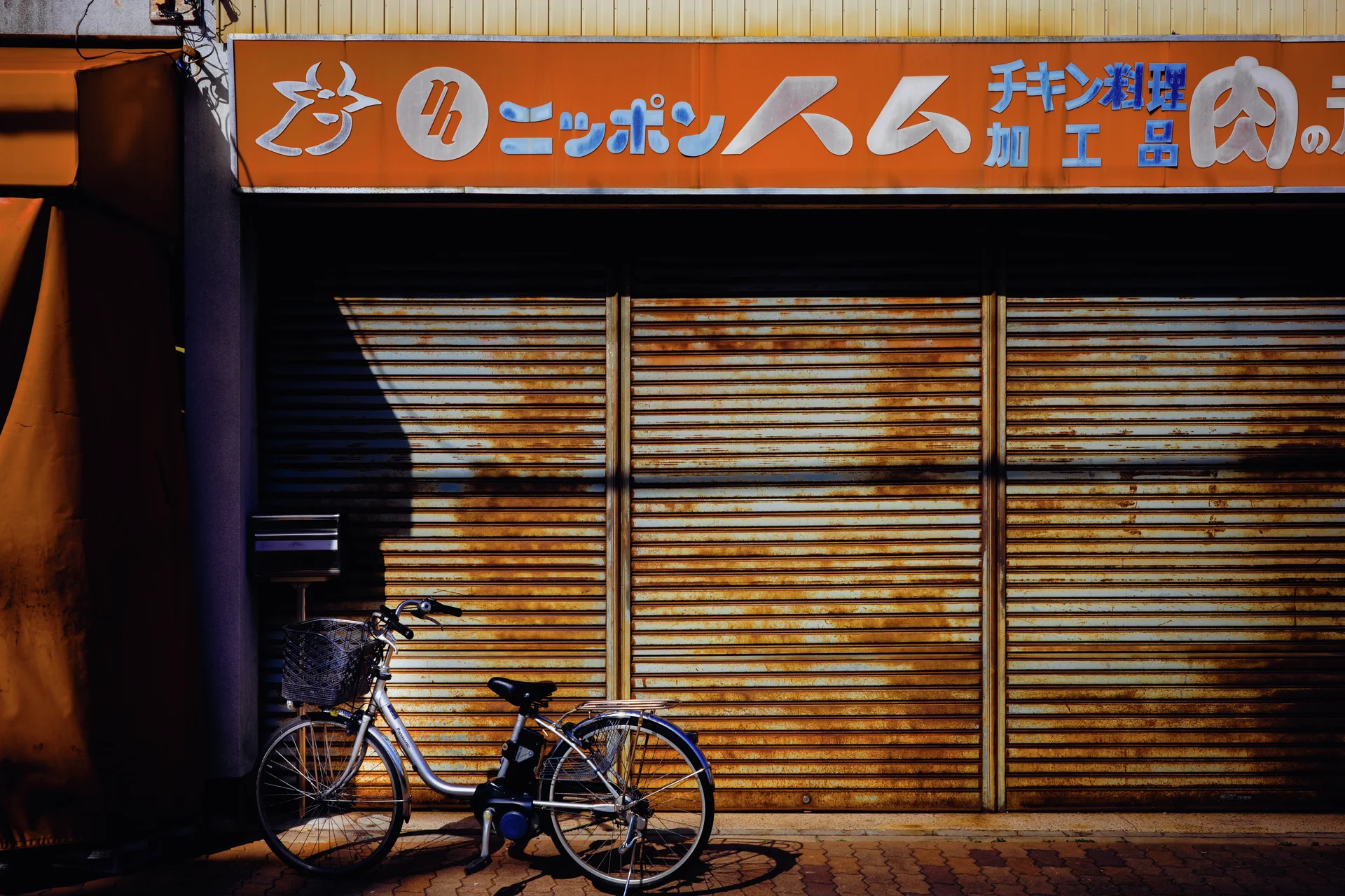 A bicycle rests before rust-streaked shutters beneath a long orange shop sign and diagonal shadows.