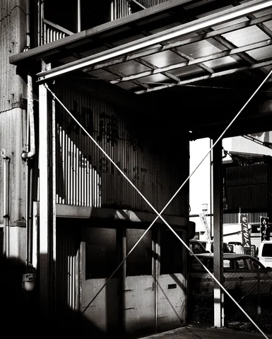 Crossed metal braces support a translucent canopy beside corrugated walls in black and white.