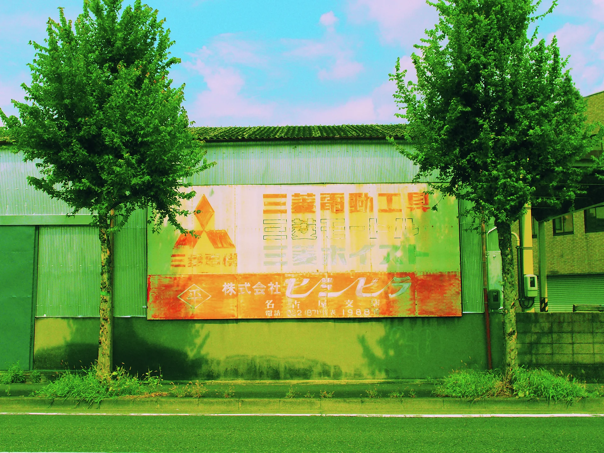 Two leafy trees flank a broad faded orange sign on a gray industrial wall.